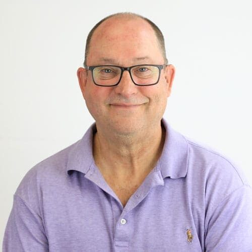 A middle-aged man with glasses and a receding hairline smiles at the camera, wearing a light purple polo shirt against a plain white background.