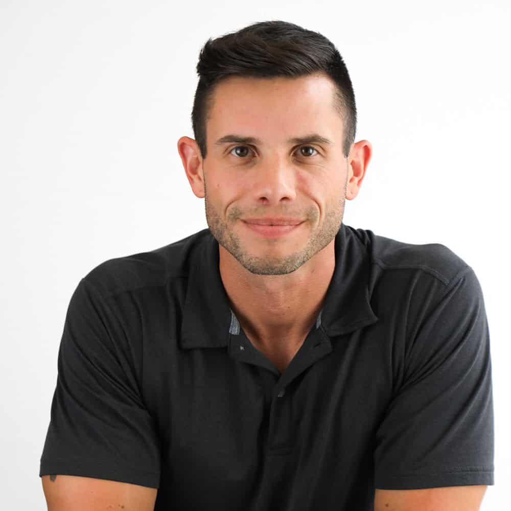 A man with short dark hair wearing a black polo shirt sits facing the camera against a plain white background.