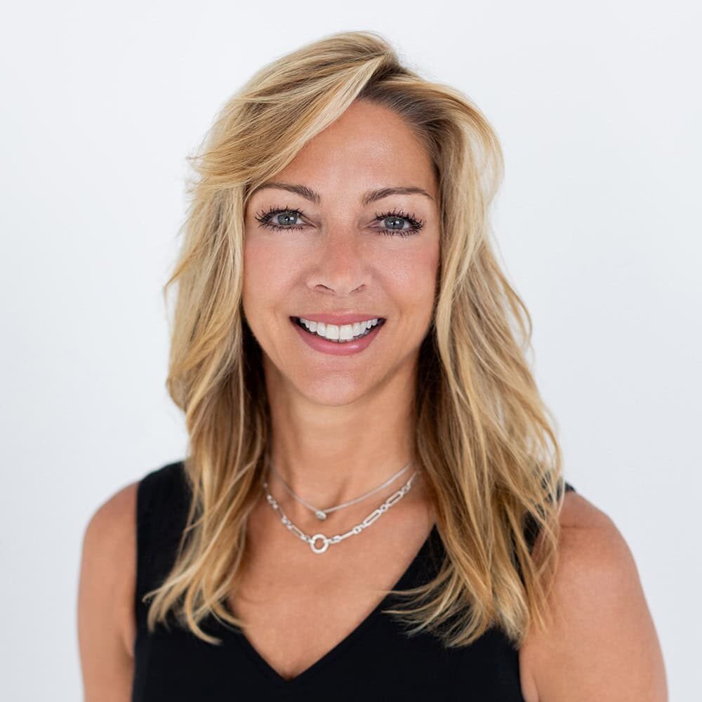 Woman with long blonde hair wearing a black sleeveless top and silver necklaces, smiling at the camera against a plain white background.