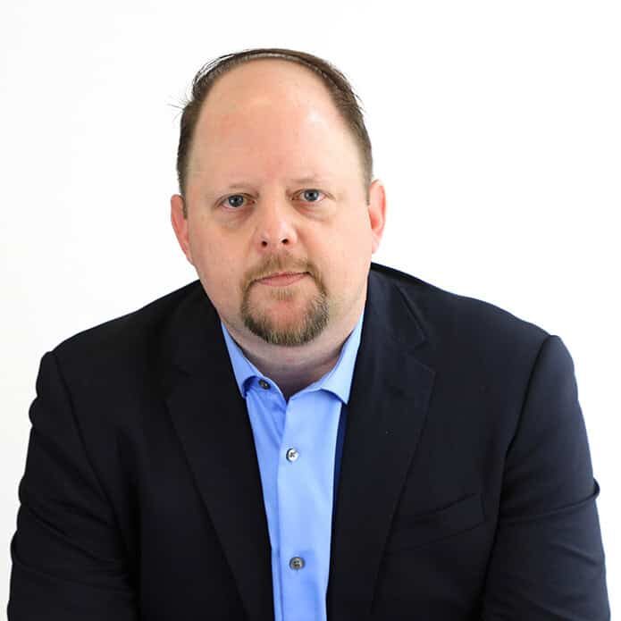 A man in a navy blazer and light blue shirt sits against a plain white background, looking directly at the camera with a neutral expression.