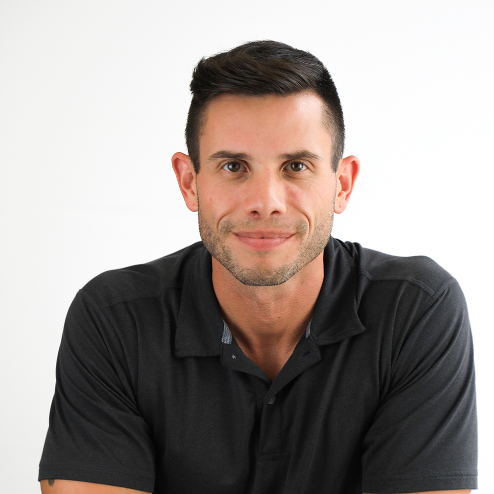 Man with short dark hair wearing a black collared shirt sits in front of a plain white background, looking directly at the camera with a slight smile.