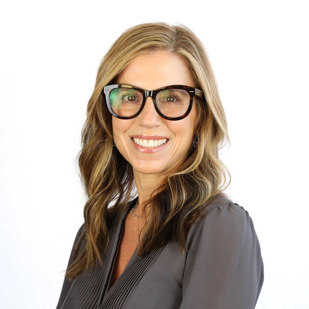 Woman with long, wavy light brown hair and black glasses, wearing a gray blouse, smiling at the camera against a plain white background.