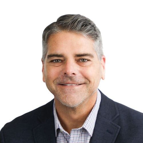 A middle-aged man with gray hair wearing a dark blazer and checked shirt smiles at the camera against a white background.